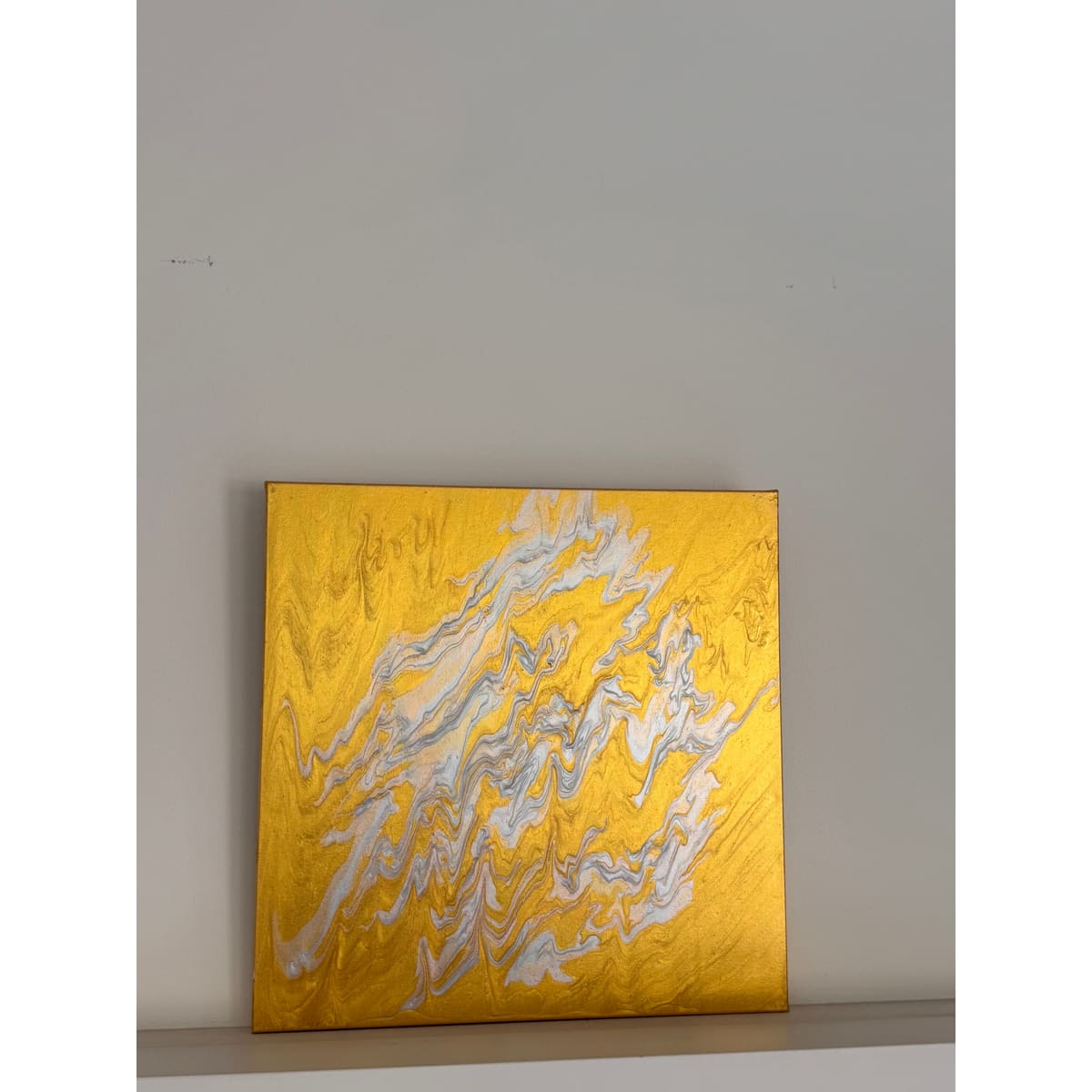 An abstract square painting with a textured gold background and flowing white and gray marbled patterns, displayed on a plain light gray wall.