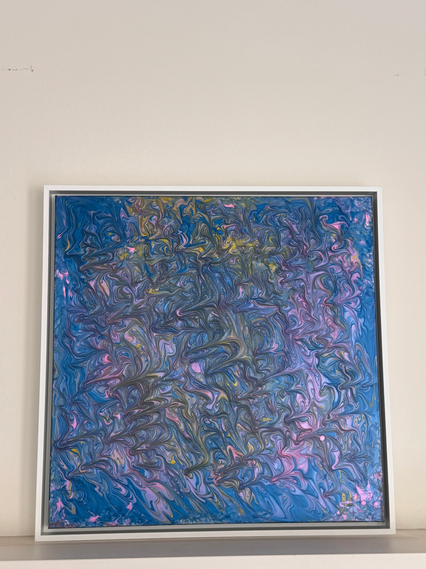 A square framed abstract painting featuring swirling, marbled textures in shades of blue, purple, pink, and hints of gold, displayed against a plain off-white wall.