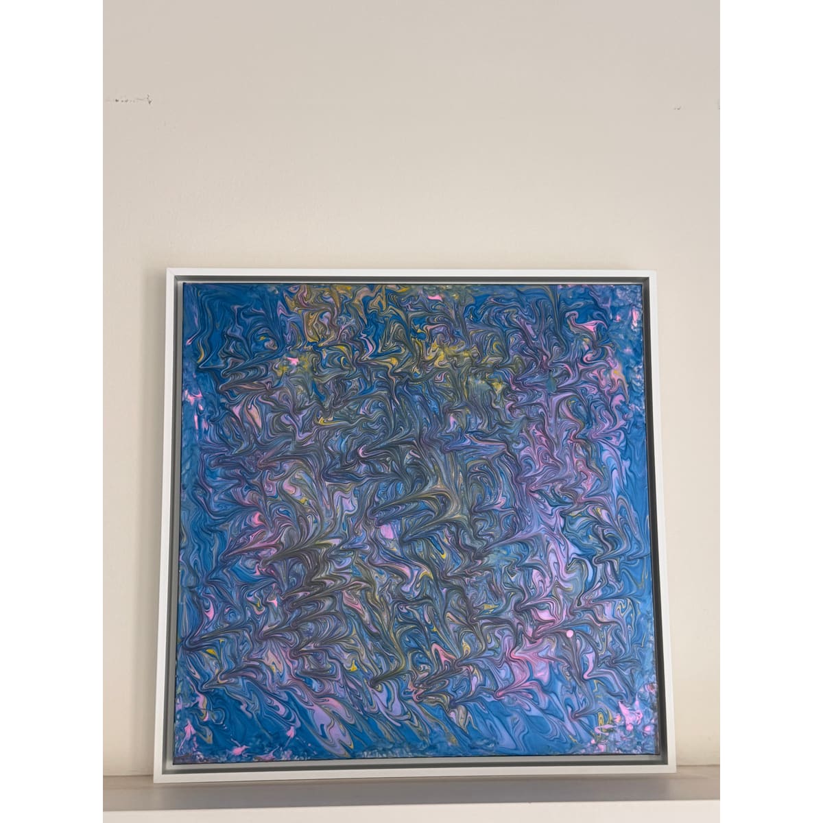 A square framed abstract painting featuring swirling, marbled textures in shades of blue, purple, pink, and hints of gold, displayed against a plain off-white wall.