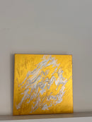 An abstract square painting with a textured gold background and flowing white and gray marbled patterns, displayed on a plain light gray wall.