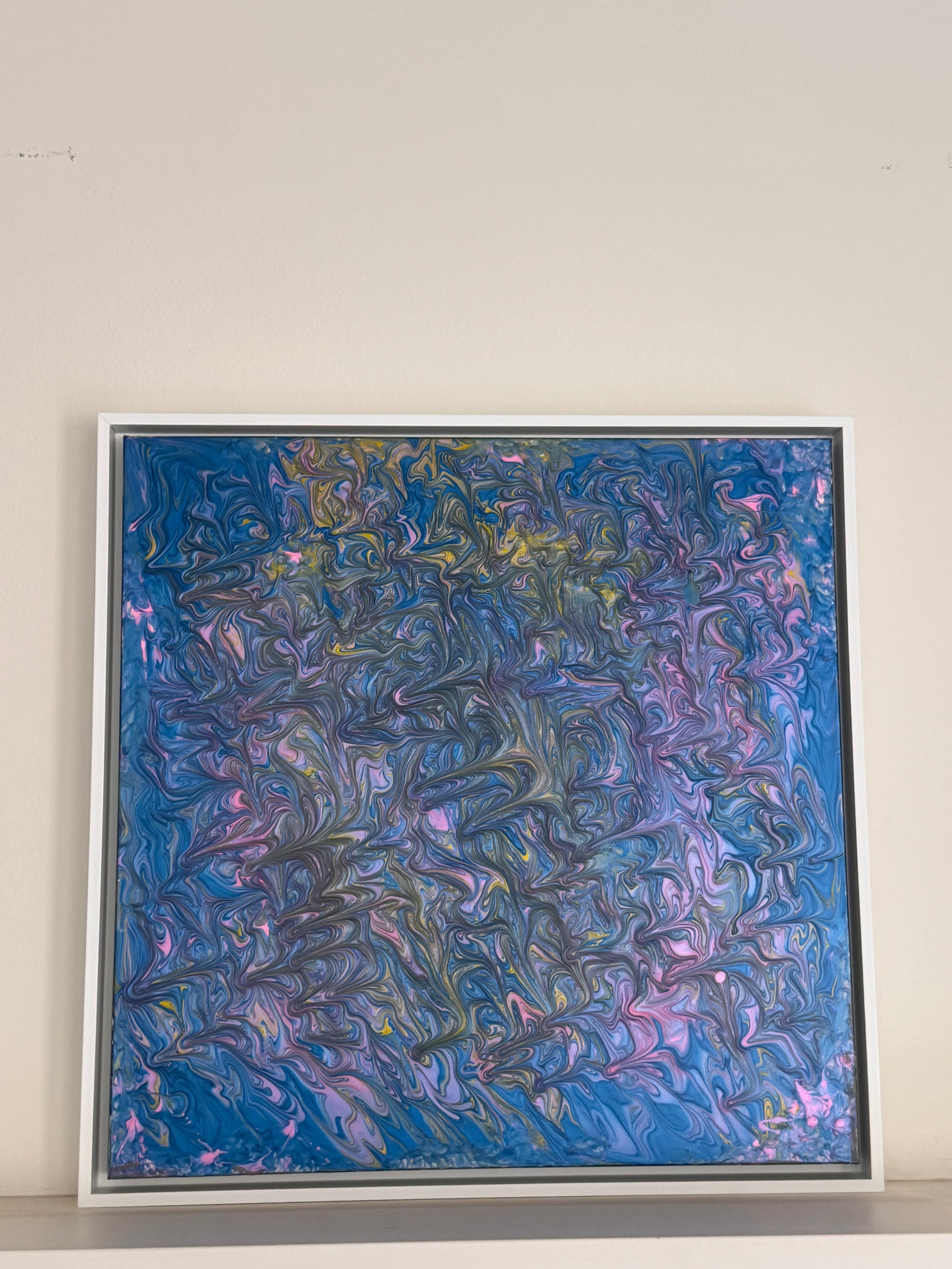 A square framed abstract painting featuring swirling, marbled textures in shades of blue, purple, pink, and hints of gold, displayed against a plain off-white wall.